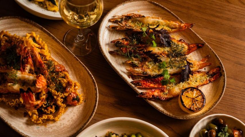 A table with grilled shrimp garnished with herbs and lemon, pasta with prawns, a bowl of mixed olives and edamame, and a glass of white wine.
